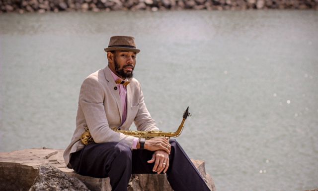 The musician Saxappeal sitting on a stone wearing a hat and the Bellis bow tie holding a saxophone in his hand